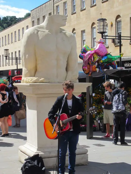 Bath, England, Stadtleben in Bath. Ein Musiker spielt vor einer Statue, der Kopf zur Hälfte fehlt. Daneben steht ein Luftballonverkäufer.